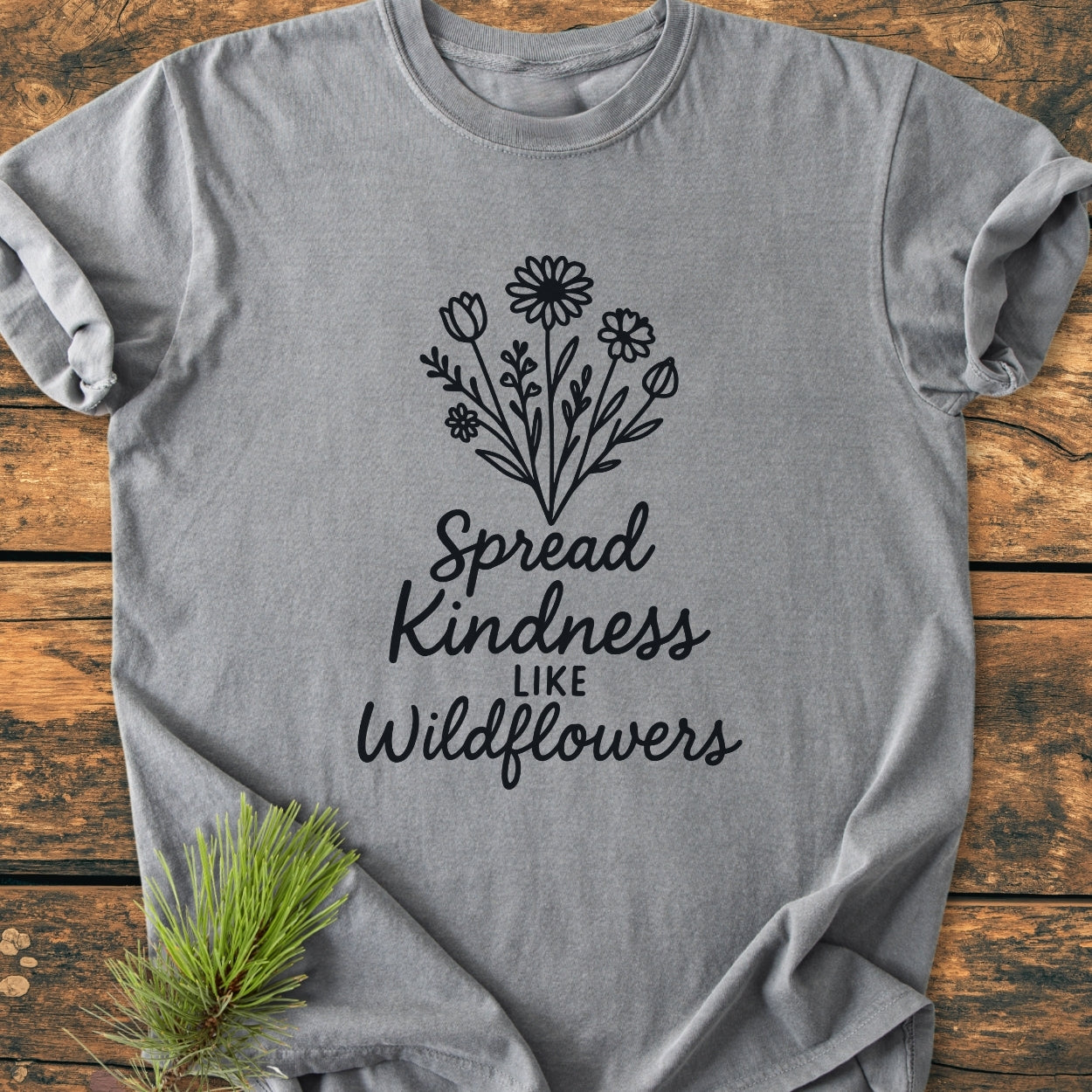Spread Kindness - Vintage Wash Tee in heather-gray, featuring black wildflowers and the phrase “Spread Kindness like Wildflowers,” laid flat on wood with a pine sprig beside it.