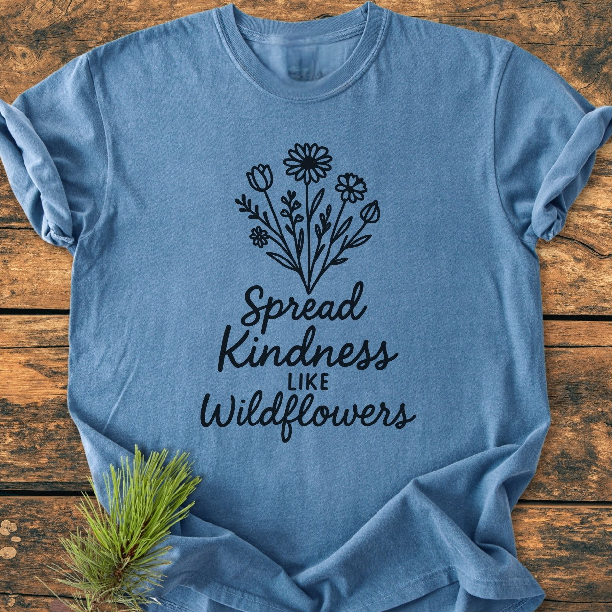 Spread Kindness - Vintage Wash Tee featuring a black wildflower bouquet and phrase on a heather-blue T-shirt, laid on rustic wood with a pine sprig for styling. Perfect for comfort and durability.