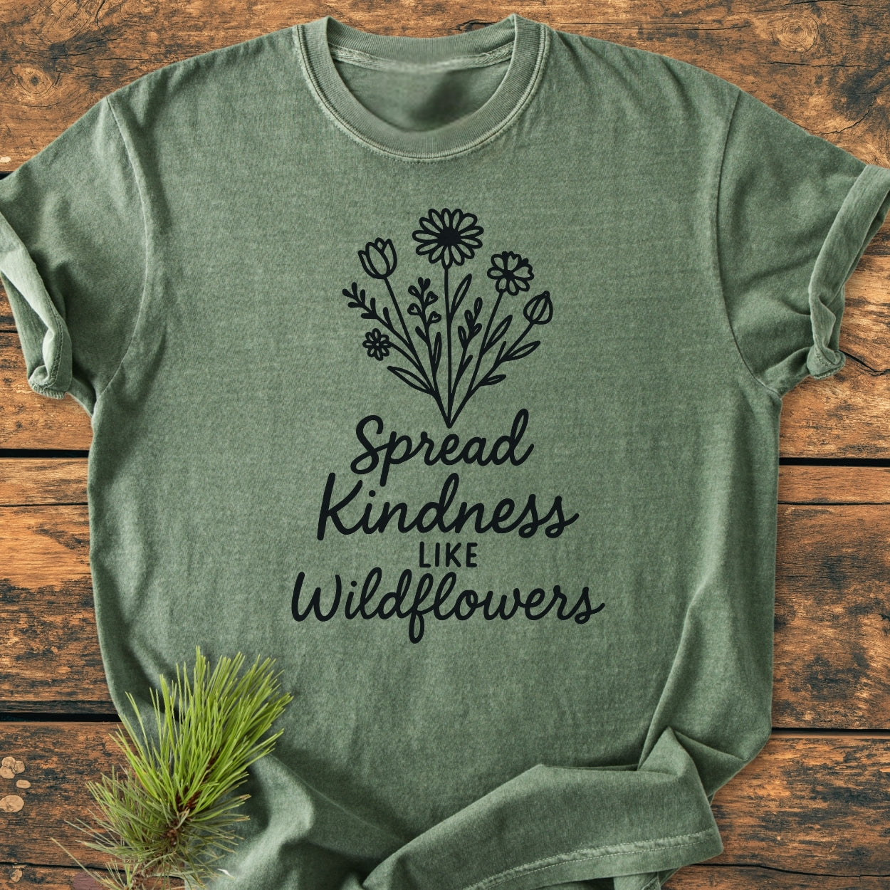 Spread Kindness - Vintage Wash Tee: A muted-green, short-sleeve T-shirt featuring black wildflowers and the phrase “Spread Kindness like Wildflowers,” laid flat on a wooden surface with a pine sprig nearby.