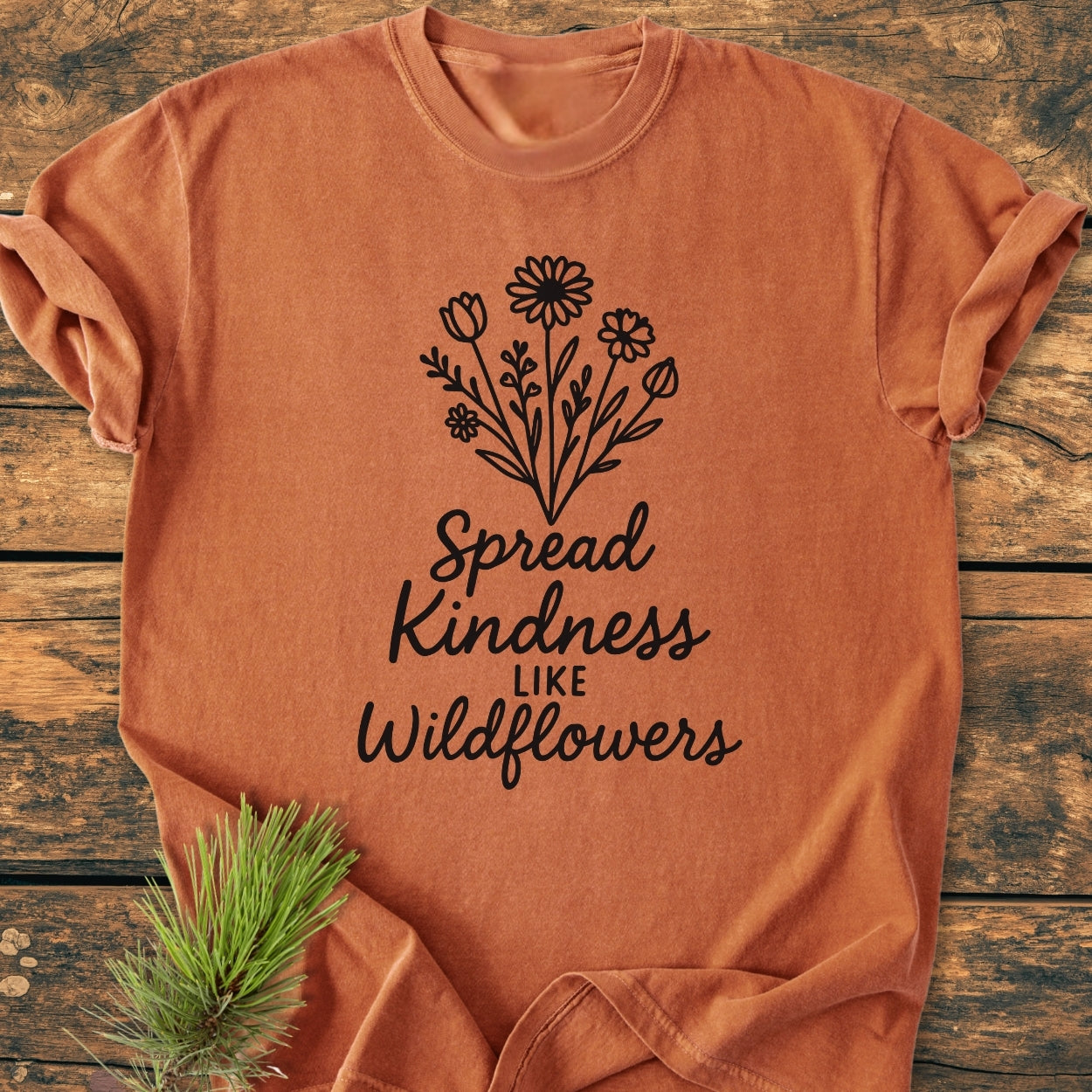 Spread Kindness - Vintage Wash Tee: A rust-orange cotton t-shirt with black wildflowers and Spread Kindness Like Wildflowers slogan, laid flat on wood with pine sprig accentuating its natural theme.