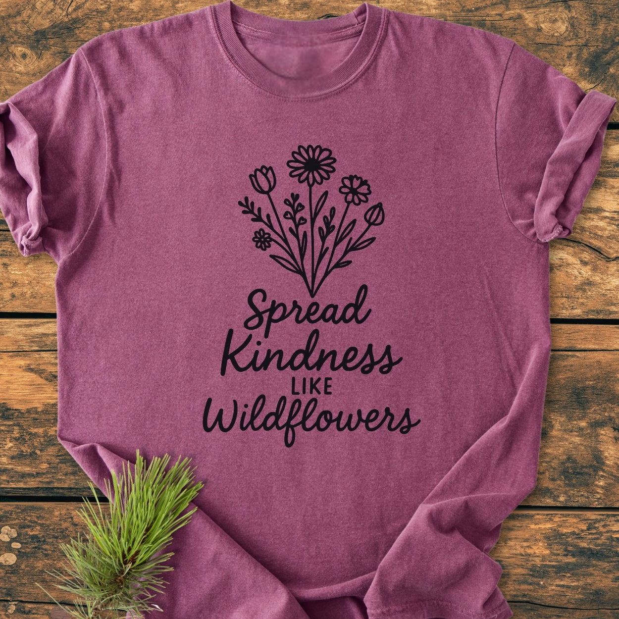 Spread Kindness - Vintage Wash Tee features a heathered plum color with wildflowers and the phrase “Spread Kindness like Wildflowers” in script, laid on rustic wood with an evergreen sprig accent.