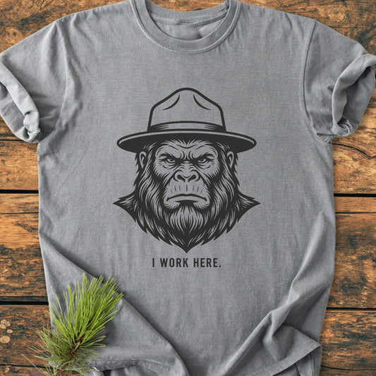 The I Work Here - Vintage Wash Tee features a stern ape illustration with a ranger hat, on heather-gray fabric, laid on wood. A pine sprig accentuates its rustic appeal.