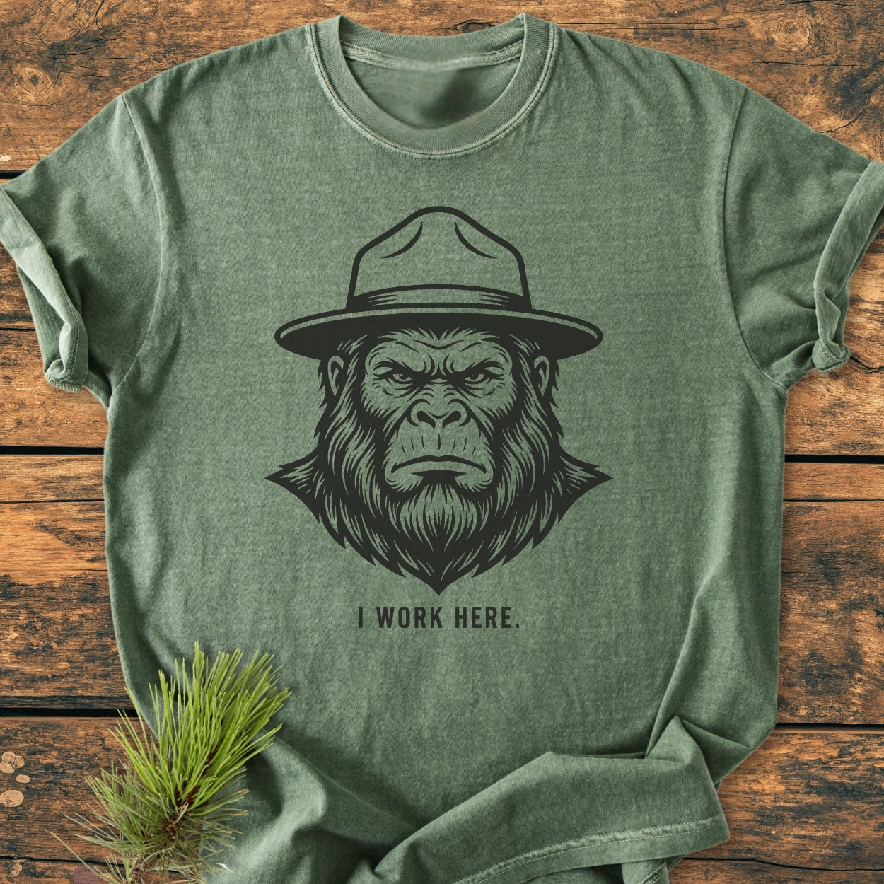 I Work Here - Vintage Wash Tee features a Sasquatch head graphic with a ranger hat on a heather-forest-green T-shirt, laid on wood, with pine needles accentuating the wilderness theme.