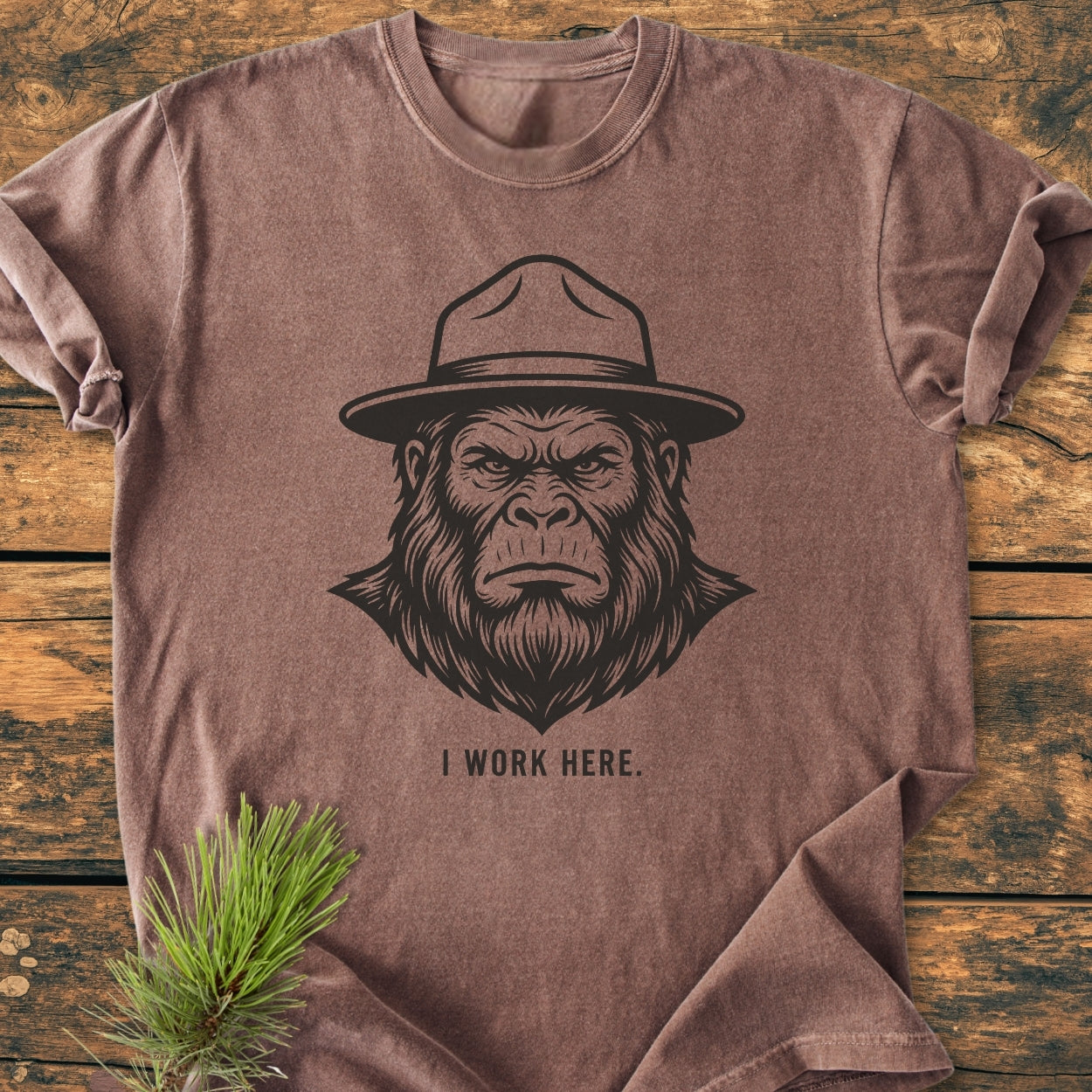 I Work Here - Vintage Wash Tee featuring a stern ape face with a ranger hat and caption. Laid on wood with a pine sprig accent, highlighting its outdoor theme.