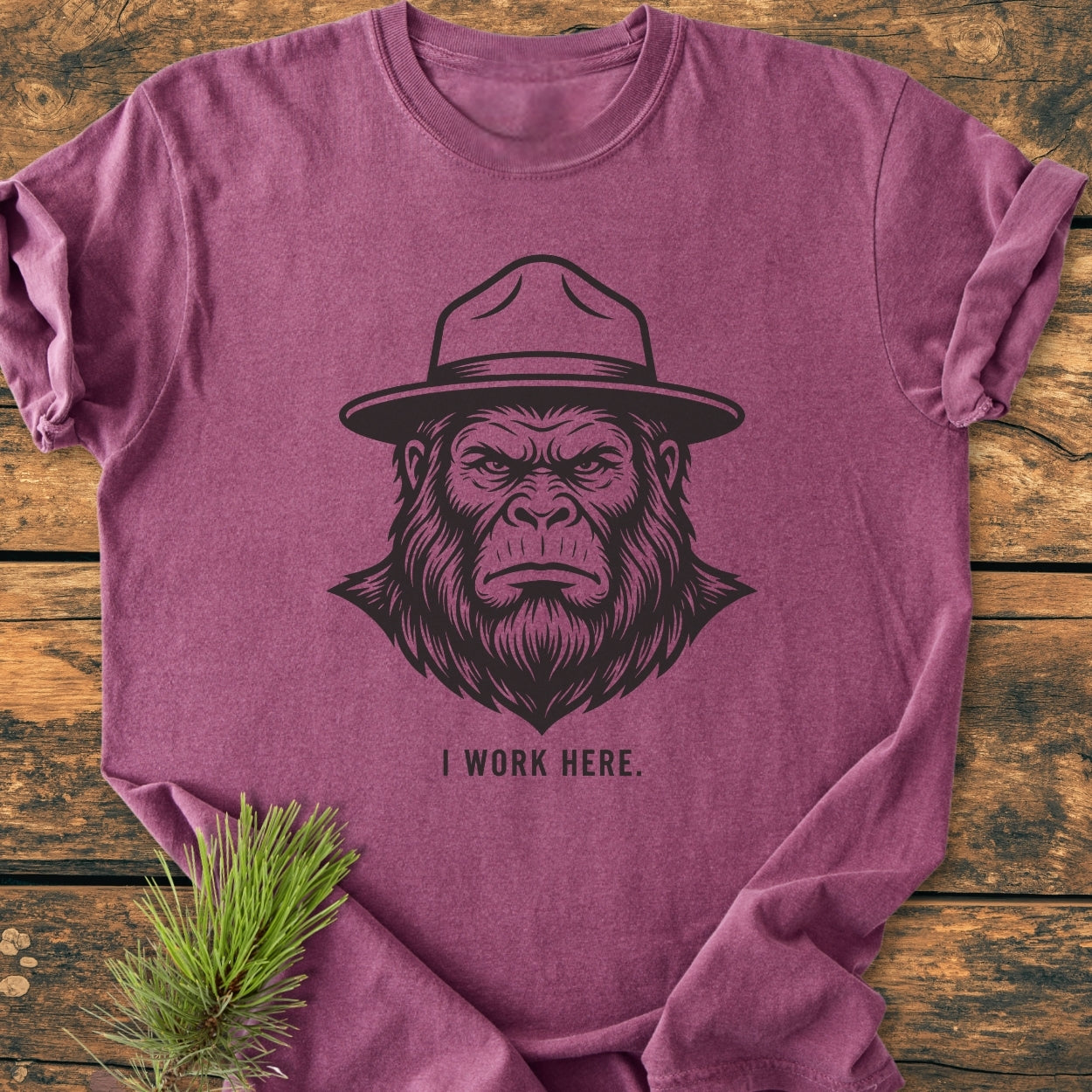Heathered purple I Work Here - Vintage Wash Tee featuring a great ape graphic with a park-ranger hat, displayed on wood, adorned with a pine branch, highlighting its cozy, relaxed fit.