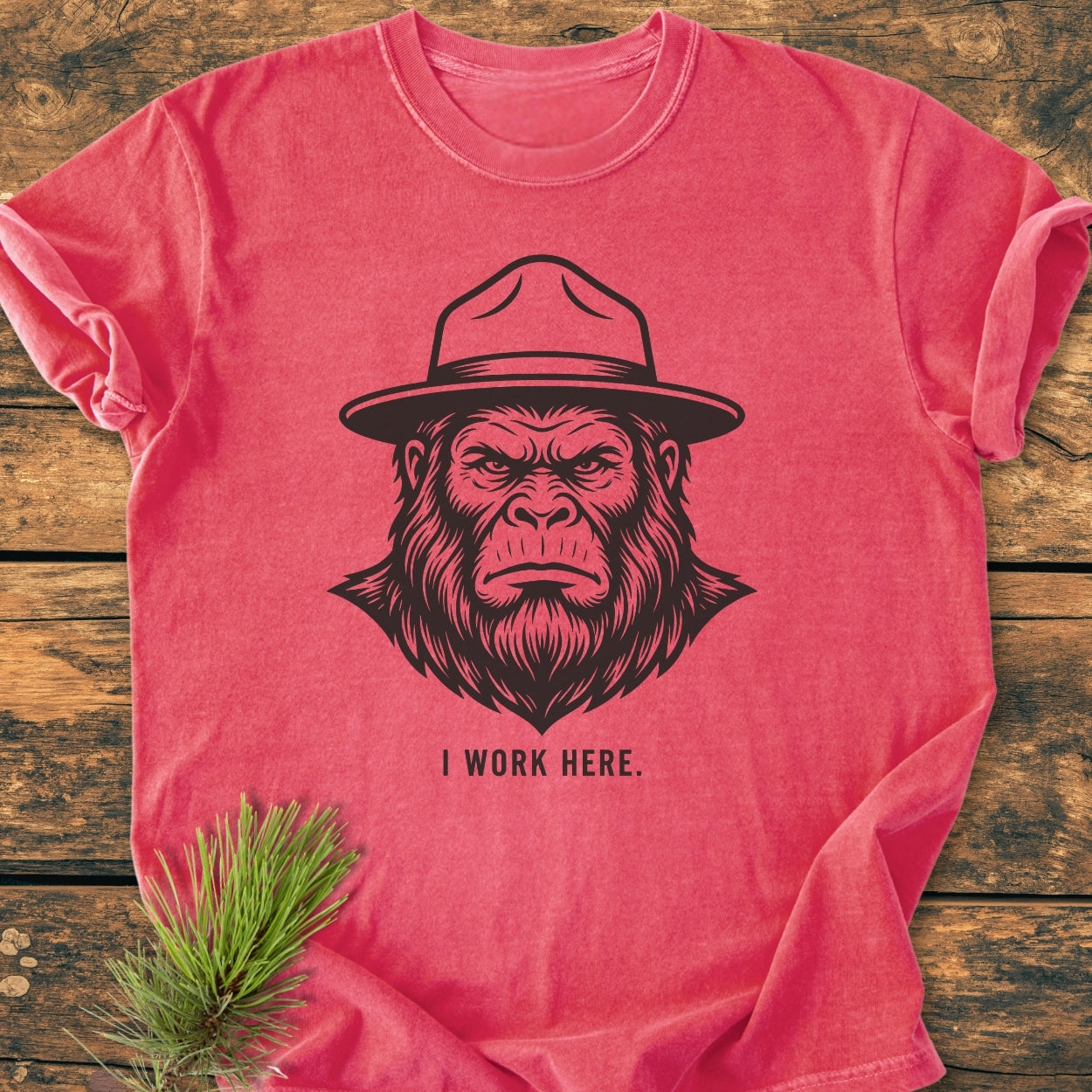 I Work Here - Vintage Wash Tee featuring a gorilla illustration with a ranger hat. Laid flat, showcasing its relaxed fit and durable stitching on a wooden surface with a pine branch accent.