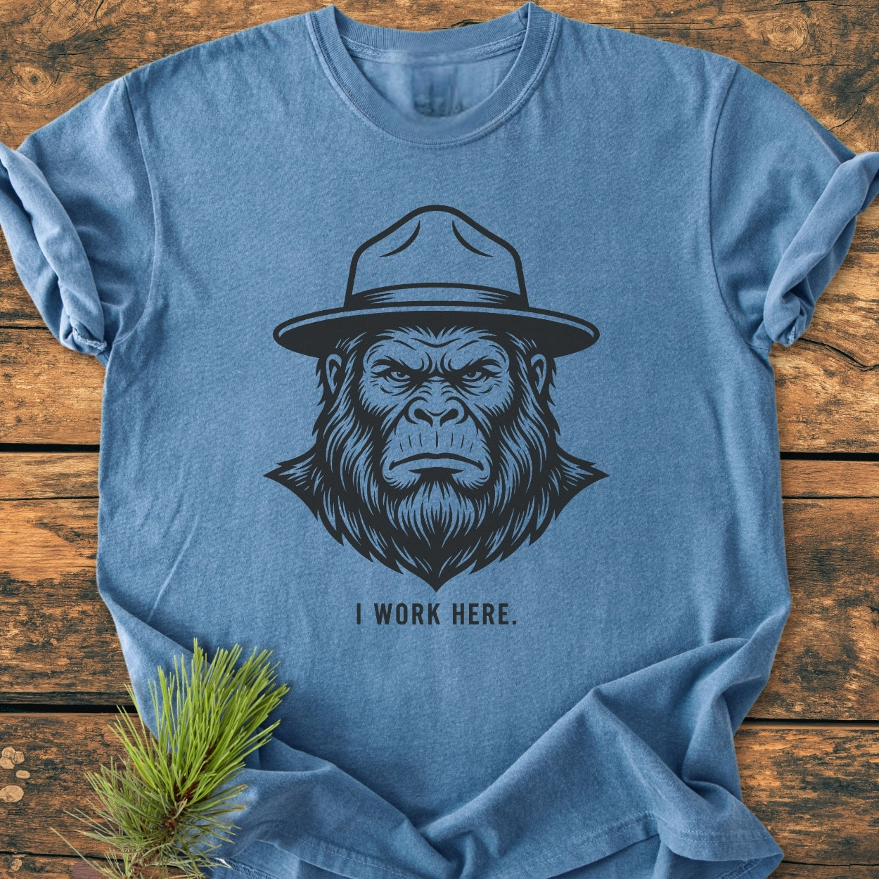 I Work Here - Vintage Wash Tee features a bold ape-like face with a ranger hat, 'I WORK HERE.' text on a heather-blue cotton fabric, laid on rustic wood with pine sprigs.