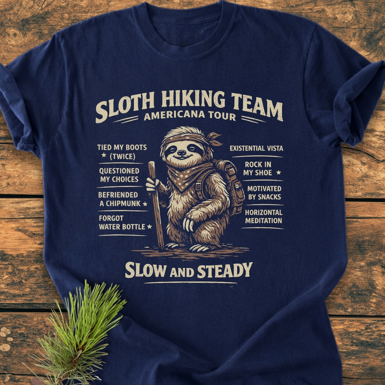 Sloth Hiking Team Tour - Vintage Wash Tee features a cartoon hiking sloth with humorous achievements, printed on a navy-blue T-shirt laid flat with a pine sprig accent.