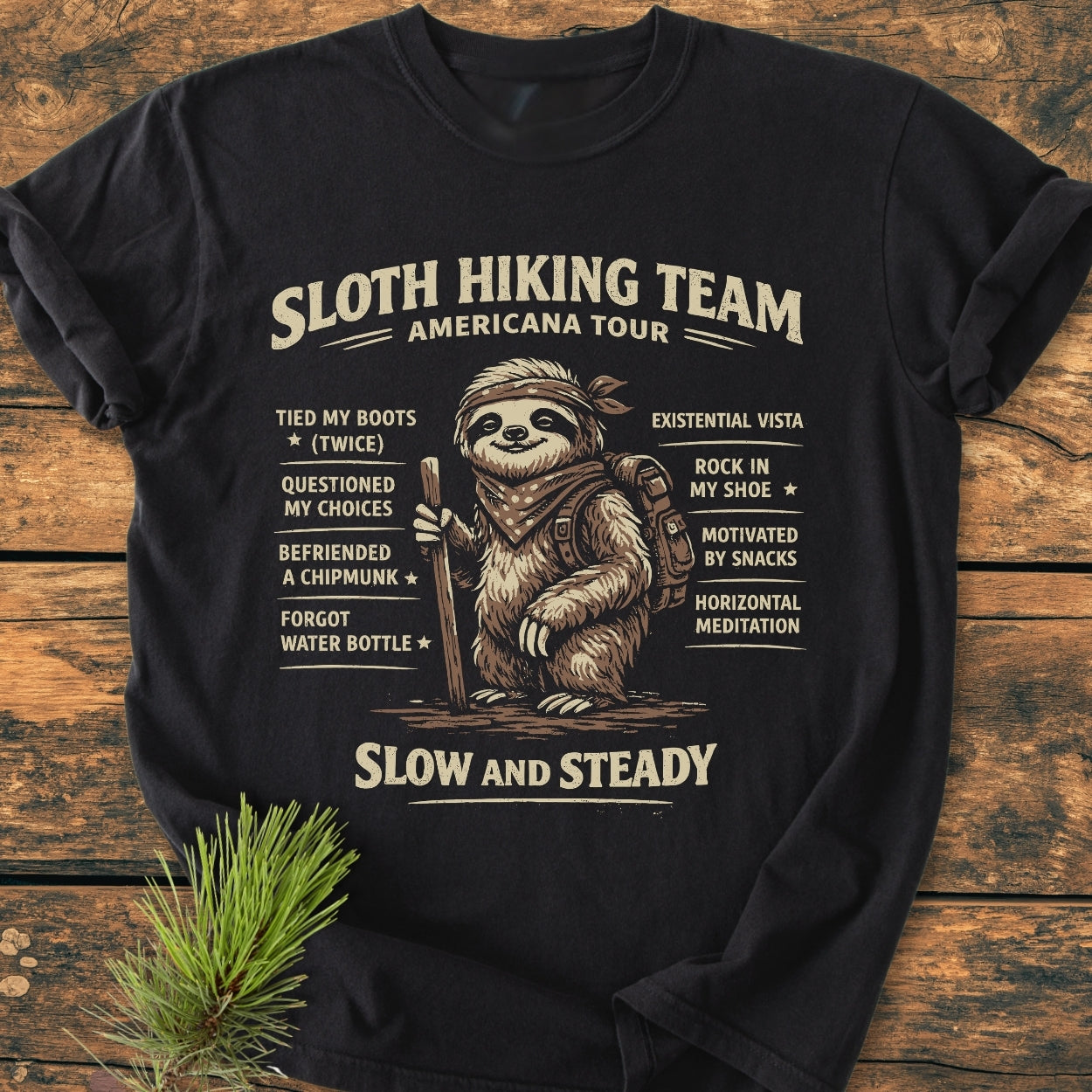 Sloth Hiking Team Tour - Vintage Wash Tee features a sloth graphic with humorous hiking achievements, laid on a wooden surface with pine sprig. The relaxed fit T-shirt offers comfort and durability.