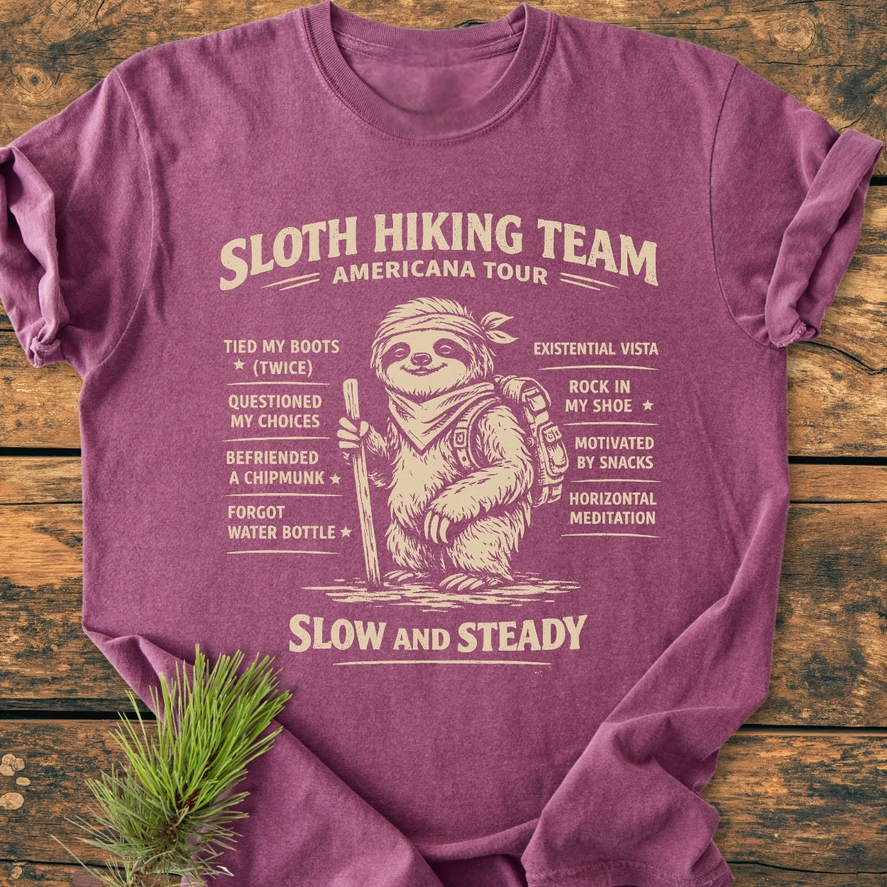 Sloth Hiking Team Tour - Vintage Wash Tee features a cartoon sloth with hiking gear and humorous checklist, emphasizing a relaxed, adventurous vibe on a maroon-purple garment-dyed T-shirt laid on wood.