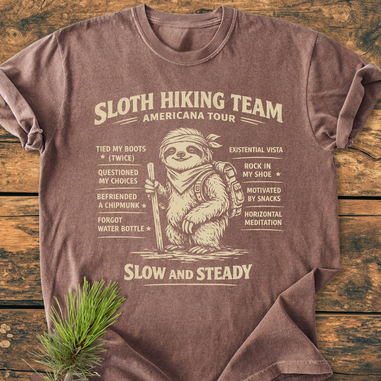 Sloth Hiking Team Tour - Vintage Wash Tee featuring a cartoon sloth graphic with hiking checklist, laid on wooden surface; showcases cozy, durable Comfort Colors 1717 garment-dyed fabric and relaxed fit.
