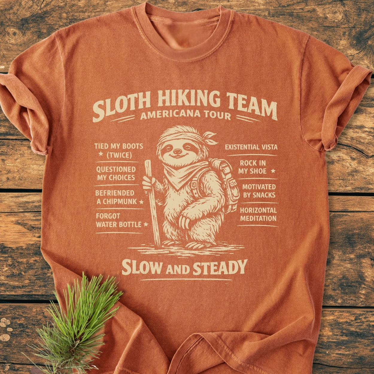 Sloth Hiking Team Tour - Vintage Wash Tee featuring a cartoon sloth with hiking gear and humorous hiking accomplishments. Laid on a wooden surface with an evergreen sprig beside it.