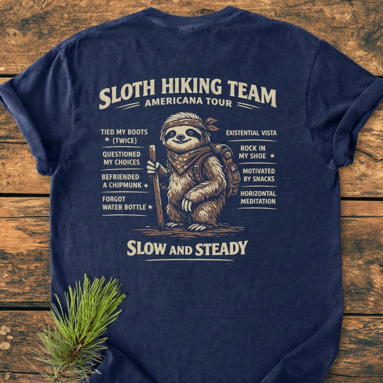 Sloth Hiking Team Tour BACK PRINT - Vintage Wash Tee featuring a sloth illustration with humorous achievements, laid on wood with a pine sprig. Made from soft, pre-shrunk, ring-spun cotton for comfort.
