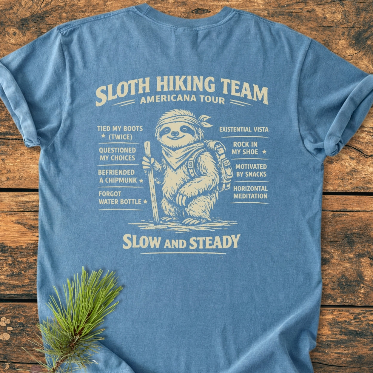 Sloth Hiking Team Tour BACK PRINT - Vintage Wash Tee: Heather-blue T-shirt featuring a playful sloth hiking design, humorous checklist, and SLOW AND STEADY. slogan, laid on a wooden surface with pine sprig accent.