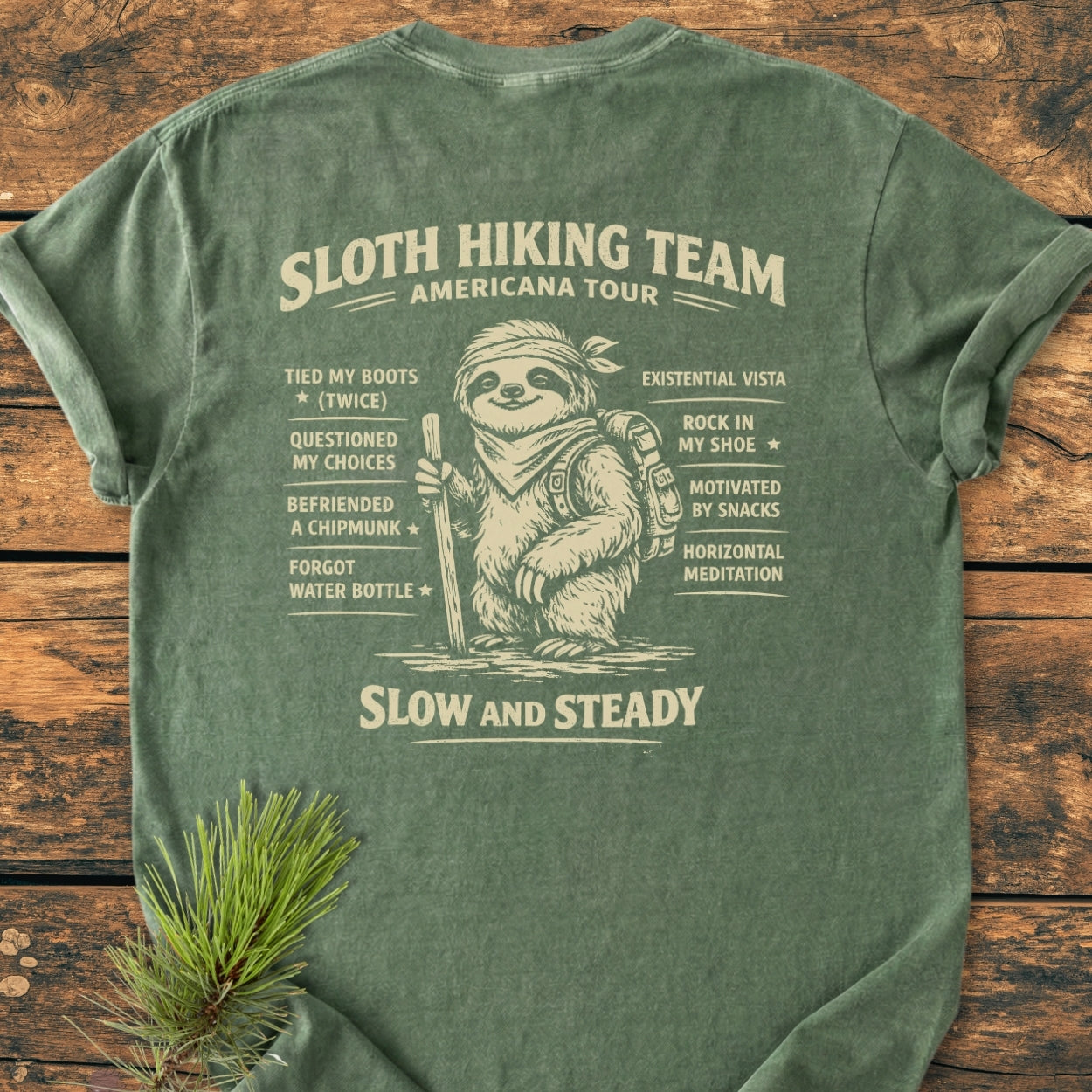 Sloth Hiking Team Tour BACK PRINT - Vintage Wash Tee features a cartoon sloth with hiking gear and humorous tour stops, printed on a green T-shirt laid on a wooden surface.