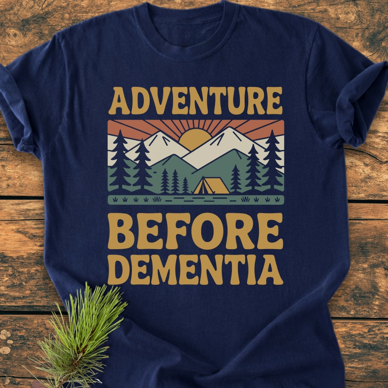 Adventure Before Dementia - Vintage Wash Tee: A navy-blue, soft-washed T-shirt featuring a retro outdoor scene with pine trees, hills, mountains, and sun, laid flat with a camping-themed pine branch beside it.