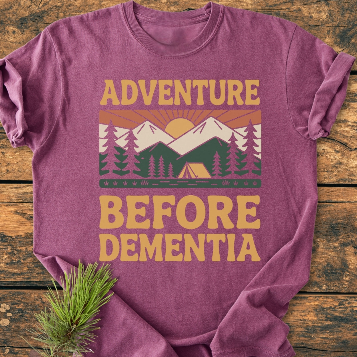 Adventure Before Dementia - Vintage Wash Tee featuring a stylized outdoor scene with mountains, pine trees, and tent, laid on wood with a pine sprig for a rustic touch.