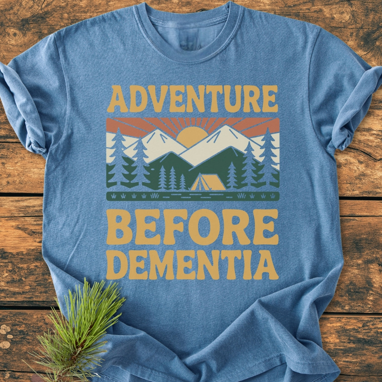 Adventure Before Dementia - Vintage Wash Tee featuring a retro sun, mountains, and campsite graphic, laid on wood, with pine needles accent. Cozy, relaxed fit, 100% ring-spun cotton.