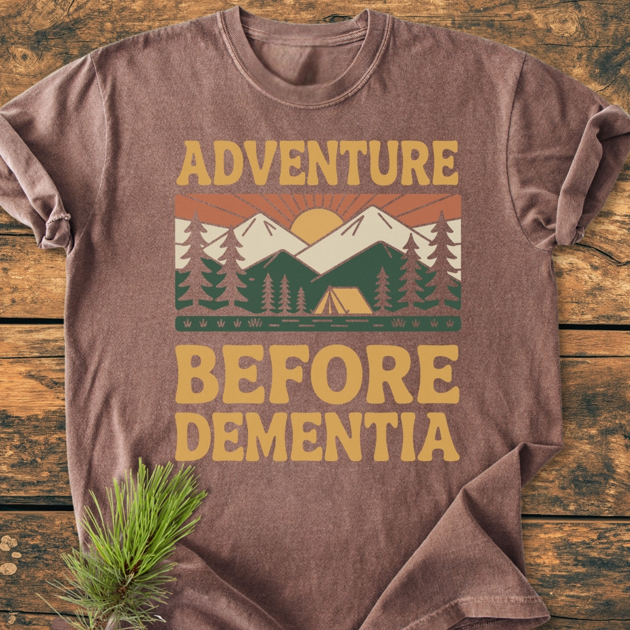 Adventure Before Dementia - Vintage Wash Tee featuring a retro sunrise over mountains and tent graphic, laid on wood. Made from soft, durable, garment-dyed 100% ring-spun cotton with a relaxed fit.