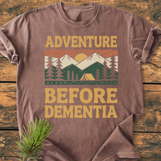 Adventure Before Dementia - Vintage Wash Tee featuring a retro sunrise over mountains and tent graphic, laid on wood. Made from soft, durable, garment-dyed 100% ring-spun cotton with a relaxed fit.