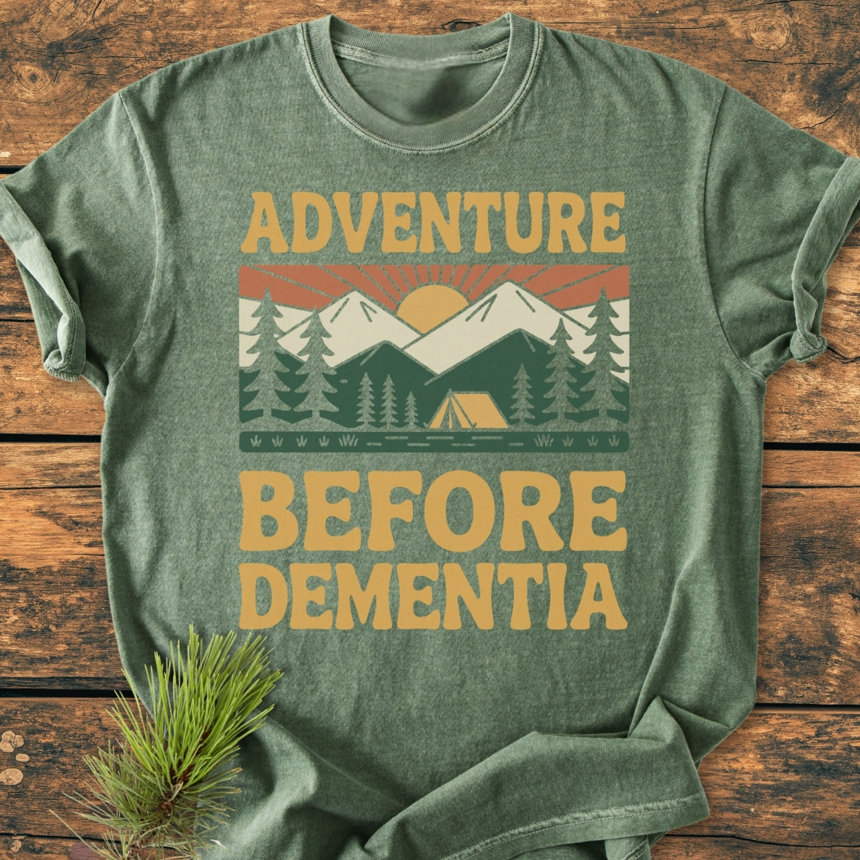 Adventure Before Dementia - Vintage Wash Tee: Heather-green crew-neck featuring a retro camping scene with a sunrise, mountains, pine trees, and a tent, printed in muted gold and forest-green tones.