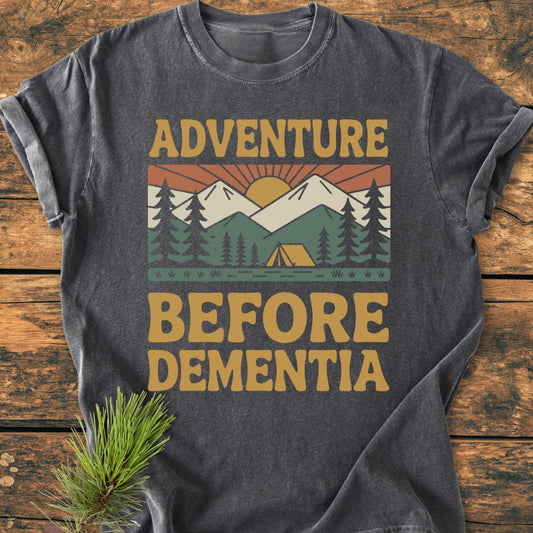 Adventure Before Dementia - Vintage Wash Tee featuring a stylized outdoor scene with pine trees, mountains, sun, and tent, printed on a dark-gray, short-sleeve T-shirt, laid flat on wood.