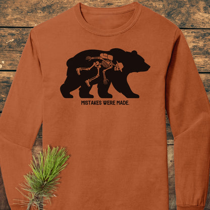 Mistakes Were Made Long-Sleeve Tee featuring a bear silhouette with a whimsical human skeleton inside. Laid on wood, adorned with a pine branch, showcasing its soft, garment-dyed, 100% ring-spun cotton construction.