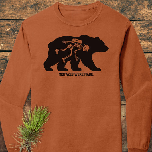 Mistakes Were Made Long-Sleeve Tee featuring a bear silhouette with a whimsical human skeleton inside. Laid on wood, adorned with a pine branch, showcasing its soft, garment-dyed, 100% ring-spun cotton construction.