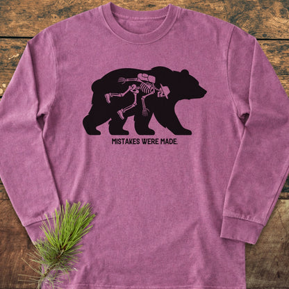 Mistakes Were Made Long-Sleeve Tee featuring a black bear silhouette with a hiker skeleton graphic, captioned below. Laid on wood, styled with a pine sprig, highlighting its relaxed, garment-dyed fit.