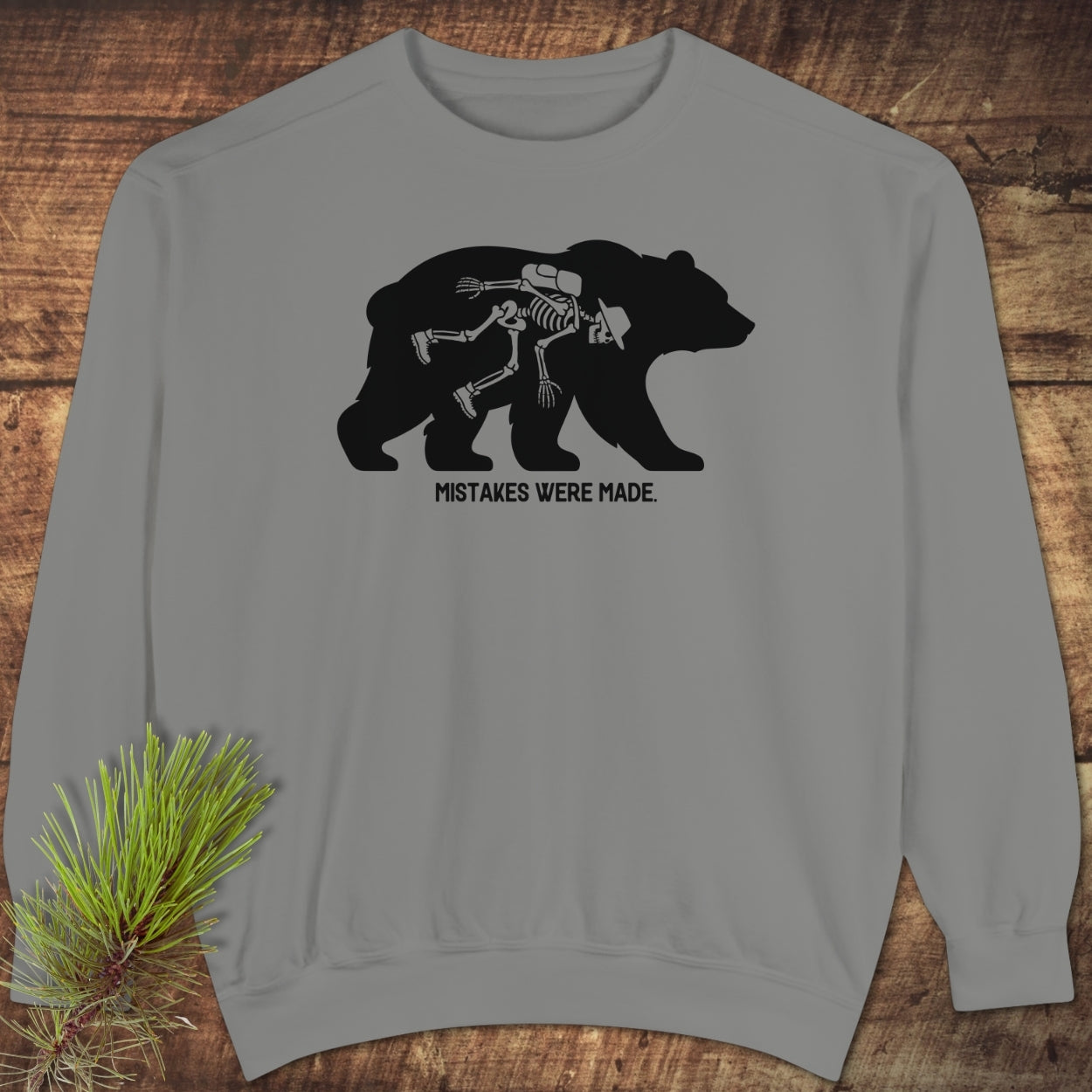 Mistakes Were Made Sweatshirt featuring a gray crewneck with a slumped skeleton inside a bear silhouette, laid on wood with pine sprig. The design humorously captures the caption “MISTAKES WERE MADE.”