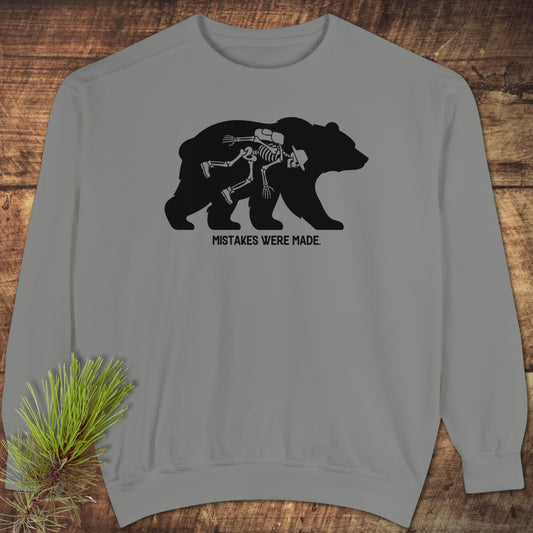 Mistakes Were Made Sweatshirt featuring a gray crewneck with a slumped skeleton inside a bear silhouette, laid on wood with pine sprig. The design humorously captures the caption “MISTAKES WERE MADE.”