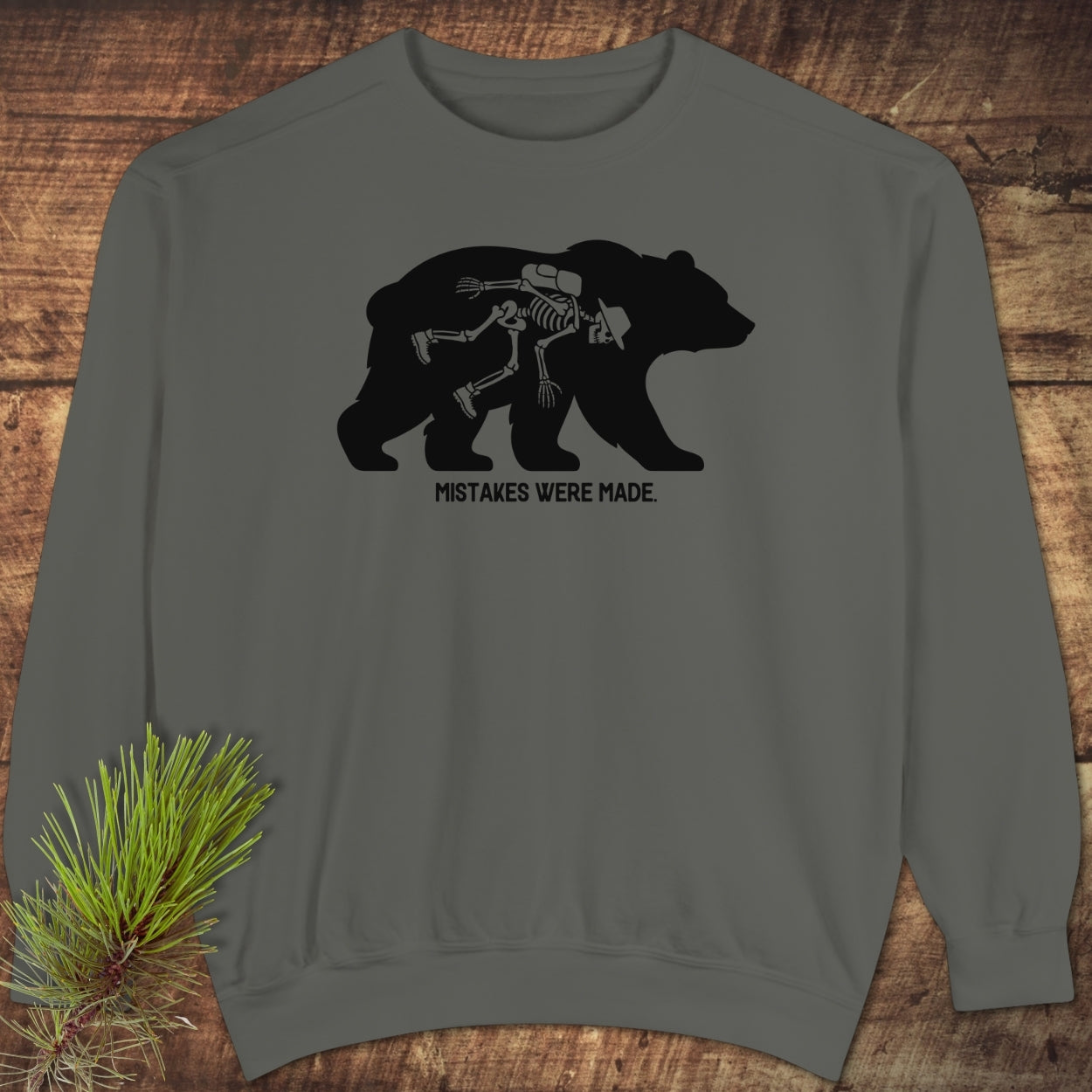 Mistakes Were Made Sweatshirt features a dark-gray crewneck with a bear silhouette and a humorous skeleton graphic. Laid on wood, it offers a relaxed fit with luxurious cotton-polyester fleece fabric.