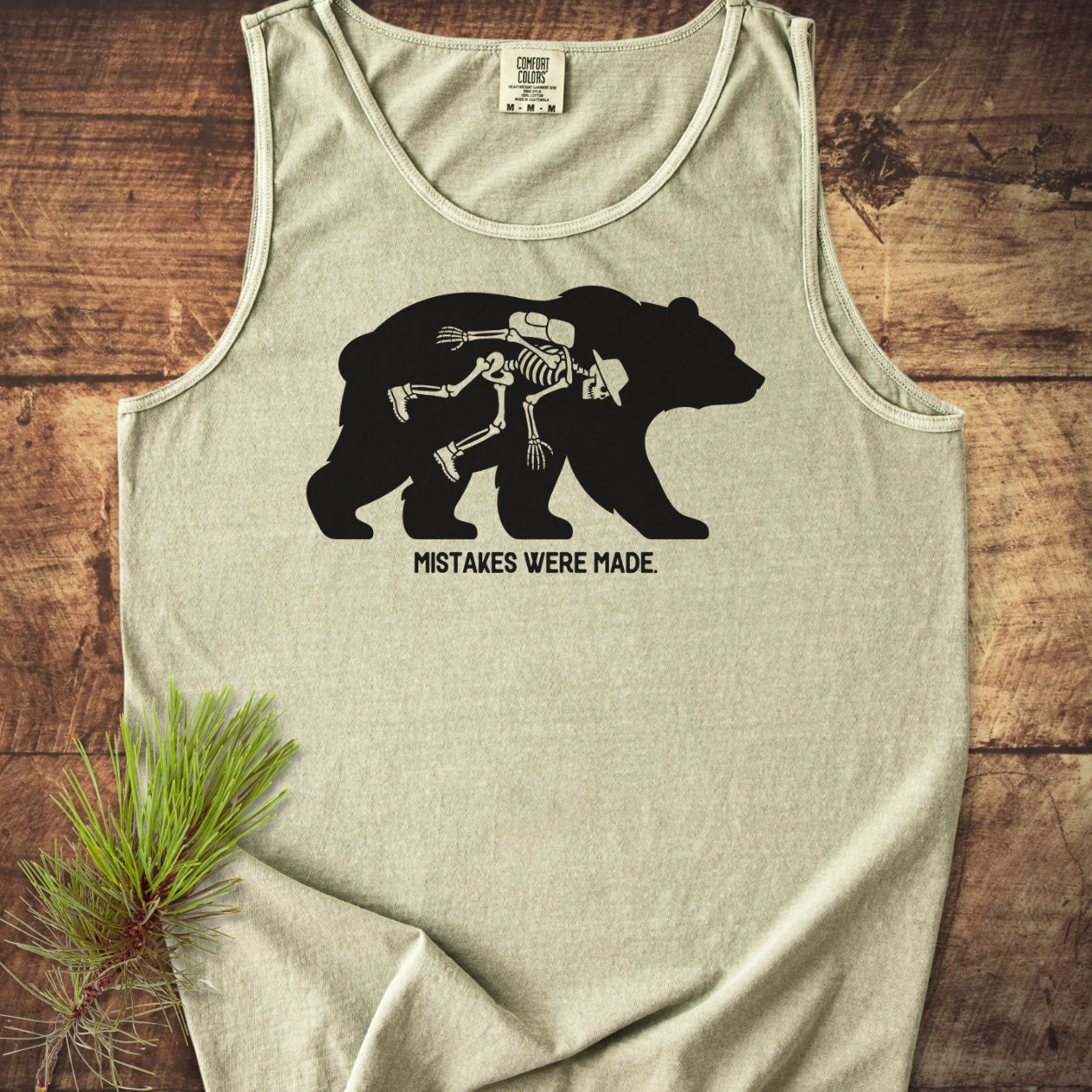 Mistakes Were Made Tank Top featuring a black bear silhouette with a hiker's skeleton outline and text below, laid on a wooden surface with a pine sprig beside it.