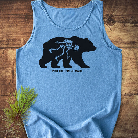 Mistakes Were Made Tank Top features a black bear graphic with a hiker’s skeleton inside, on a heather-blue fabric. Laid on wood with a pine sprig, emphasizing its outdoor-inspired design.