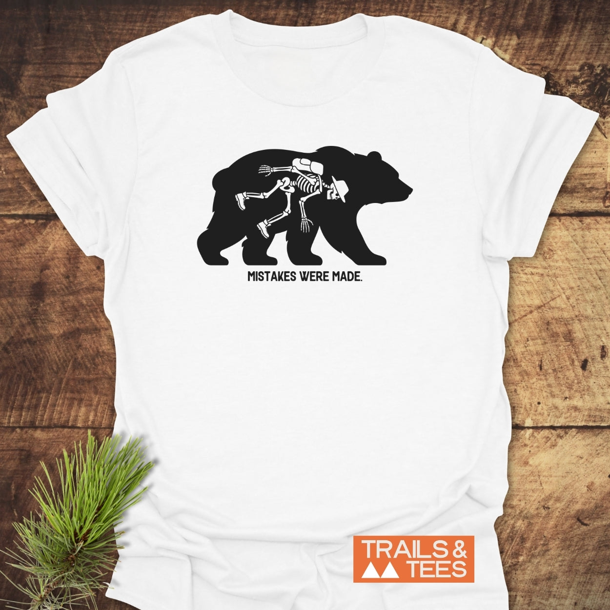 Mistakes Were Made T-Shirt featuring a bear silhouette with a human skeleton in hiking gear inside. Caption below reads “MISTAKES WERE MADE.” Includes Trails & Tees logo. Laid on wooden surface.
