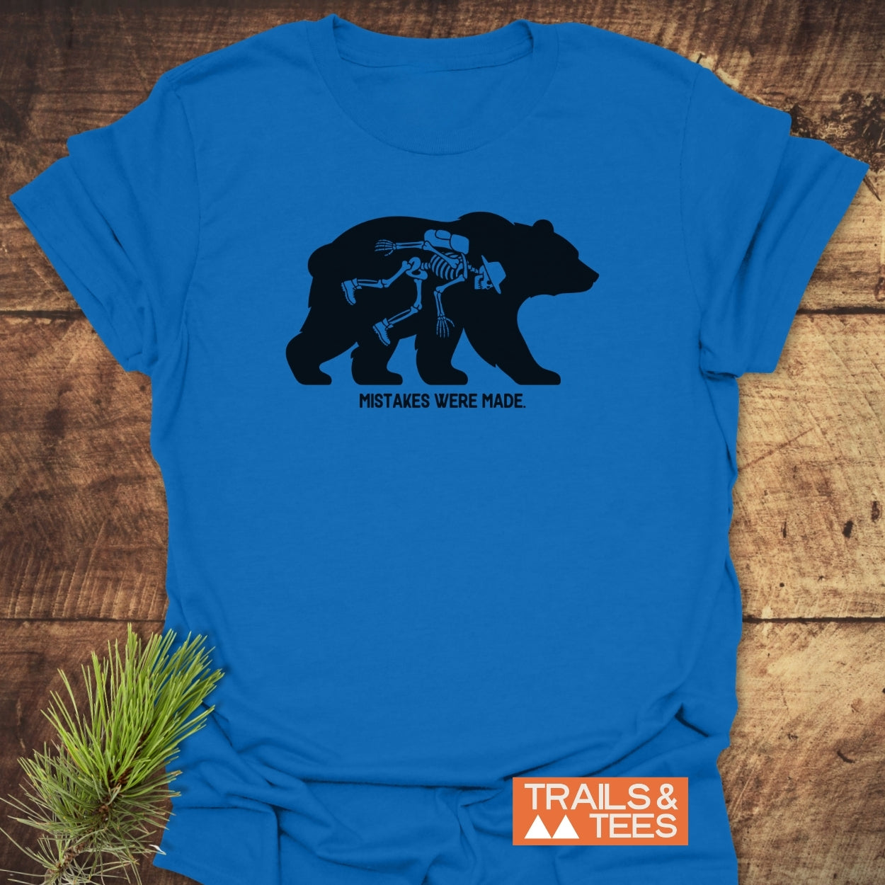 Mistakes Were Made T-Shirt features a bear graphic with a hiker's skeleton, highlighting outdoor humor. Laid on wood, it symbolizes adventure and comfort, crafted from soft ring-spun cotton.
