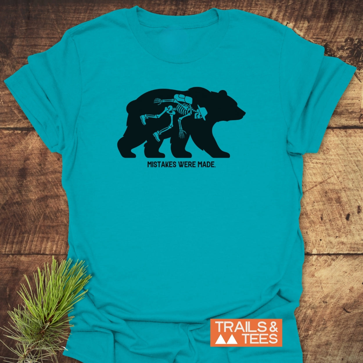 Mistakes Were Made T-Shirt featuring a bear silhouette with a hiker's skeleton inside, printed in black on a teal shirt. Includes TRAILS & TEES logo, emphasizing outdoor adventure and comfort.