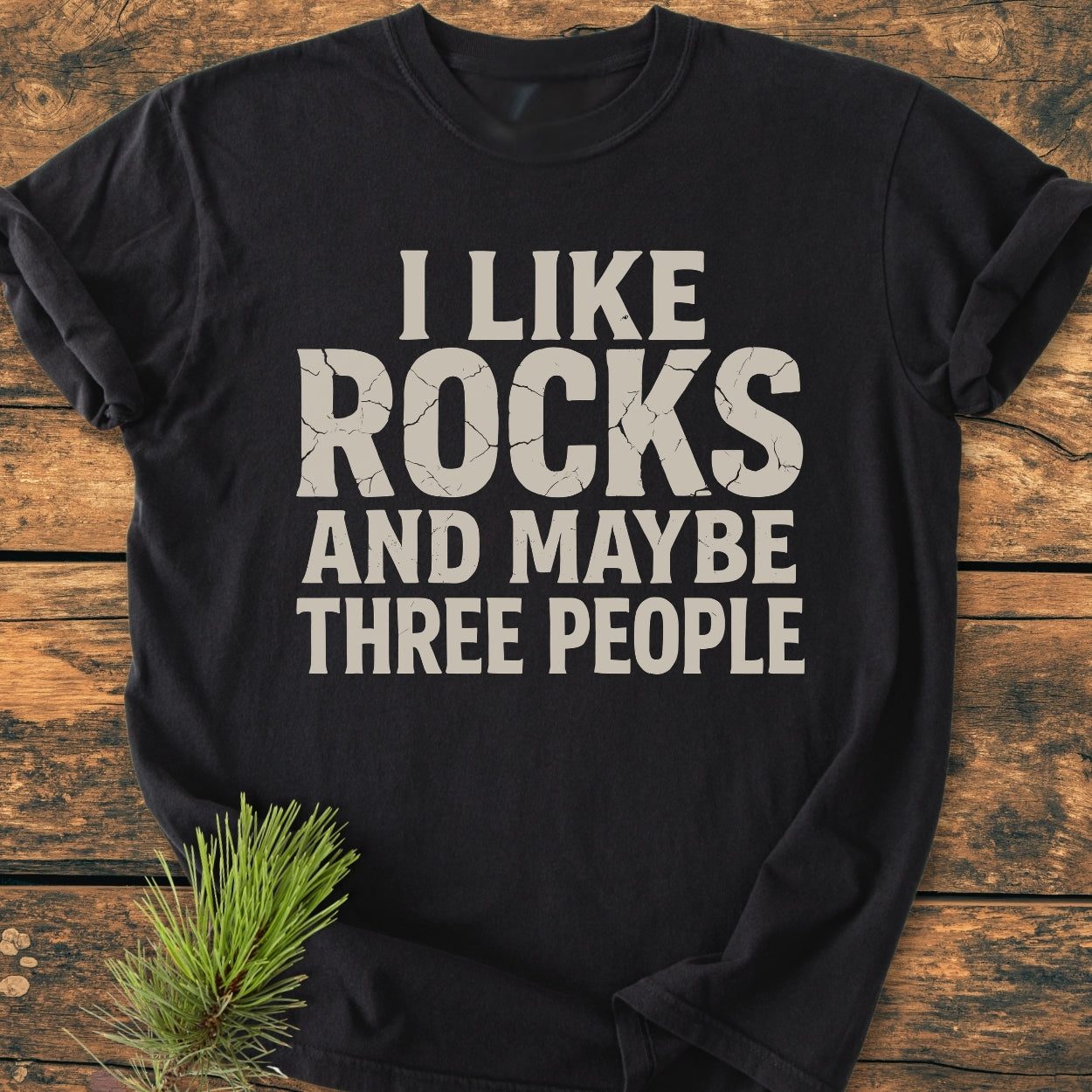 I Like Rocks - Vintage Wash Tee displayed on a wooden surface, features a cracked-letter slogan. Made of pre-shrunk, garment-dyed cotton, offering a relaxed fit, durable stitching, and tubular shape.