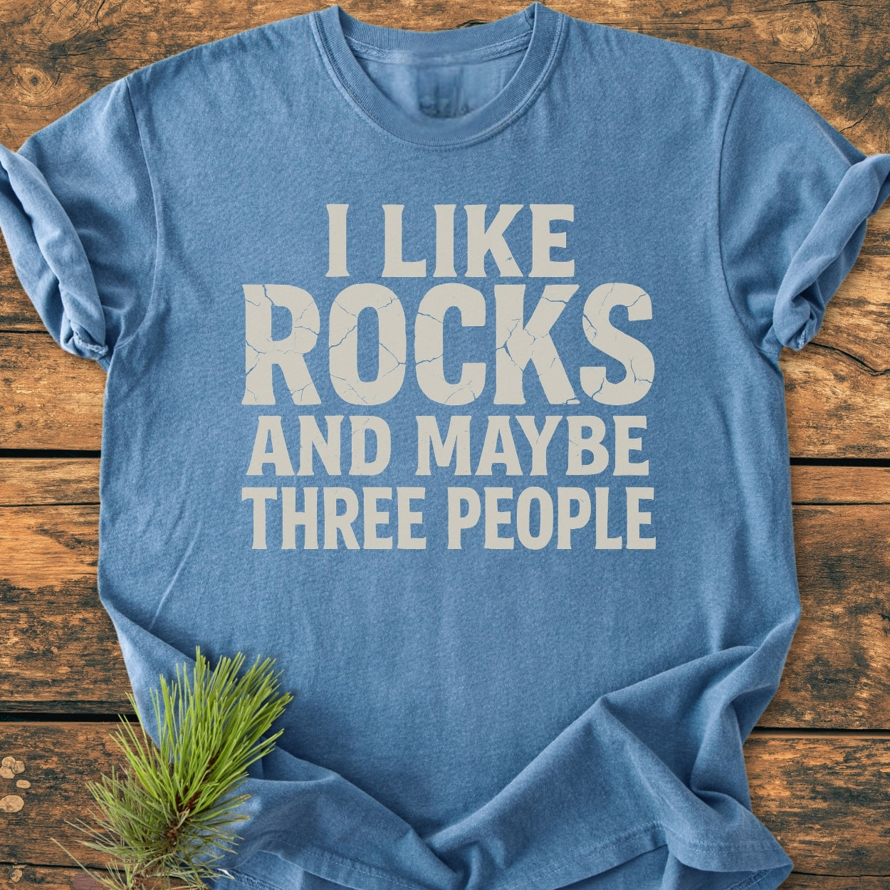 Heather-blue I Like Rocks - Vintage Wash Tee laid flat, featuring cracked-style lettering. Styled with a pine sprig on a wooden surface, showcasing its relaxed fit and soft-washed, durable fabric.