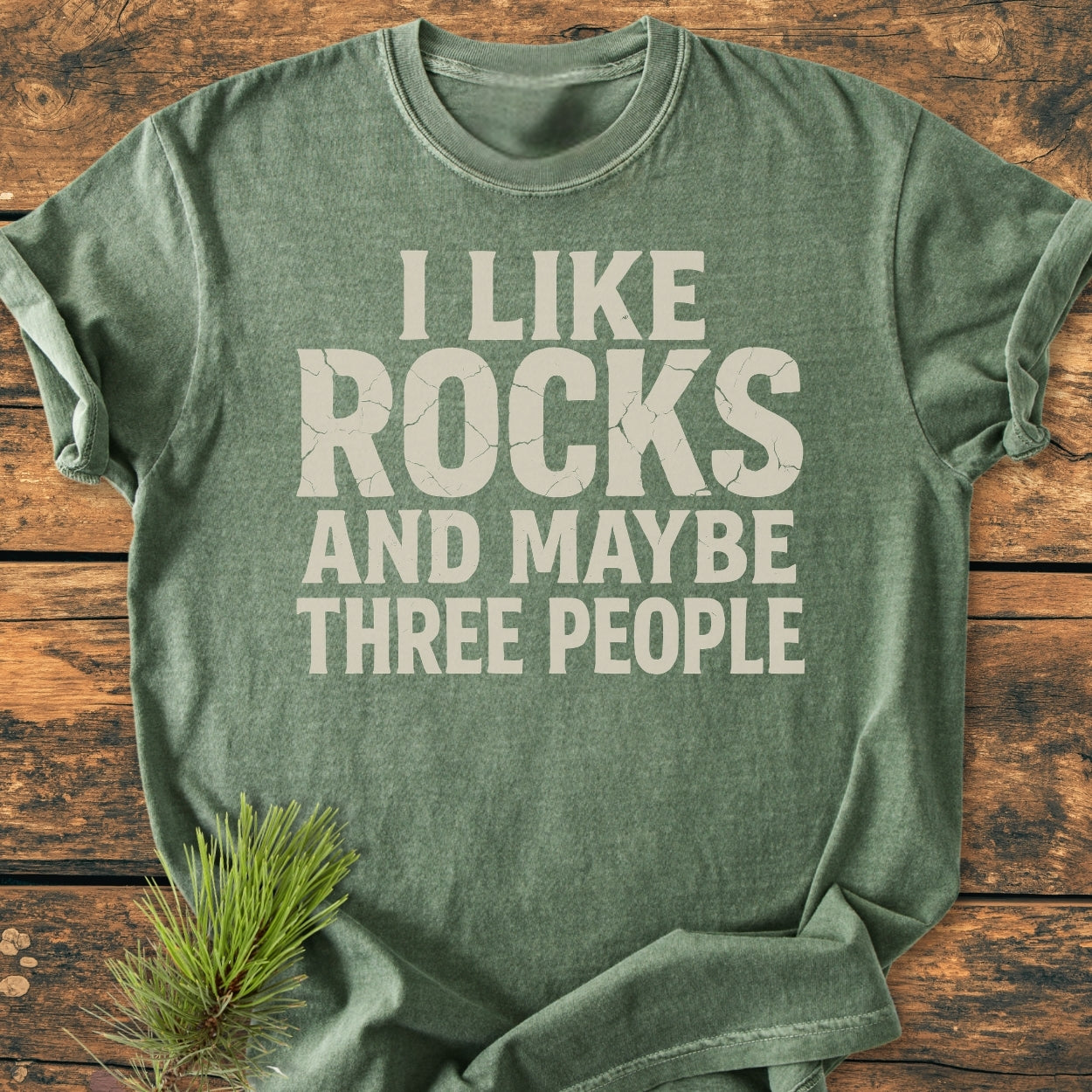 Heather-green I Like Rocks - Vintage Wash Tee with off-white slogan on rustic wood, featuring soft-washed, garment-dyed fabric, relaxed fit, and durable stitching, with a pine sprig accent nearby.
