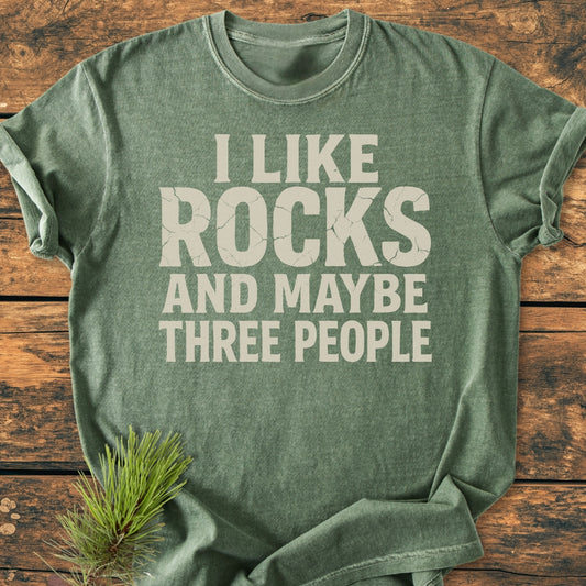 Heather-green I Like Rocks - Vintage Wash Tee with off-white slogan on rustic wood, featuring soft-washed, garment-dyed fabric, relaxed fit, and durable stitching, with a pine sprig accent nearby.