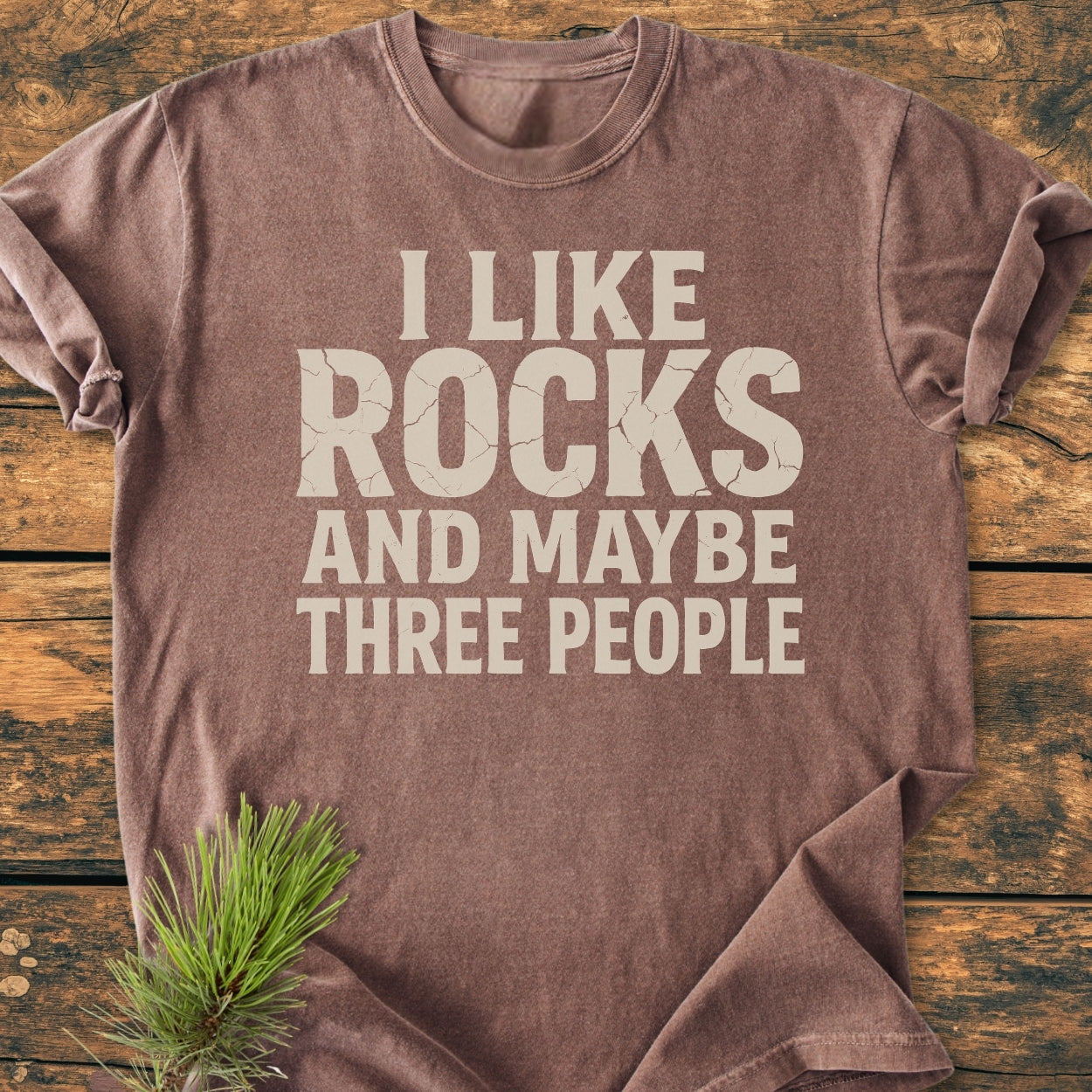 I Like Rocks - Vintage Wash Tee laid on wood, showcasing its slogan in pale print. The soft, garment-dyed fabric and relaxed fit highlight its cozy appeal, complemented by durable, double-needle stitching.