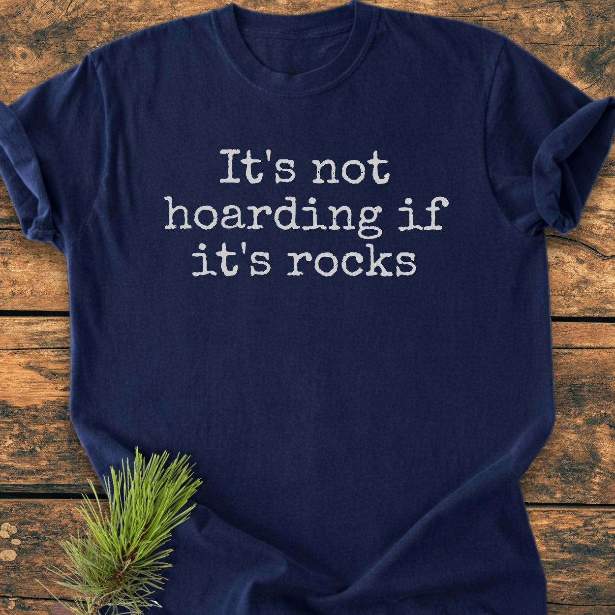 It's Not Hoarding If It's Rocks - Vintage Wash Tee displayed on a wooden surface with pine sprig; navy blue, typewriter-style slogan, soft-washed fabric, relaxed fit, durable double-needle stitching, tubular shape retention.