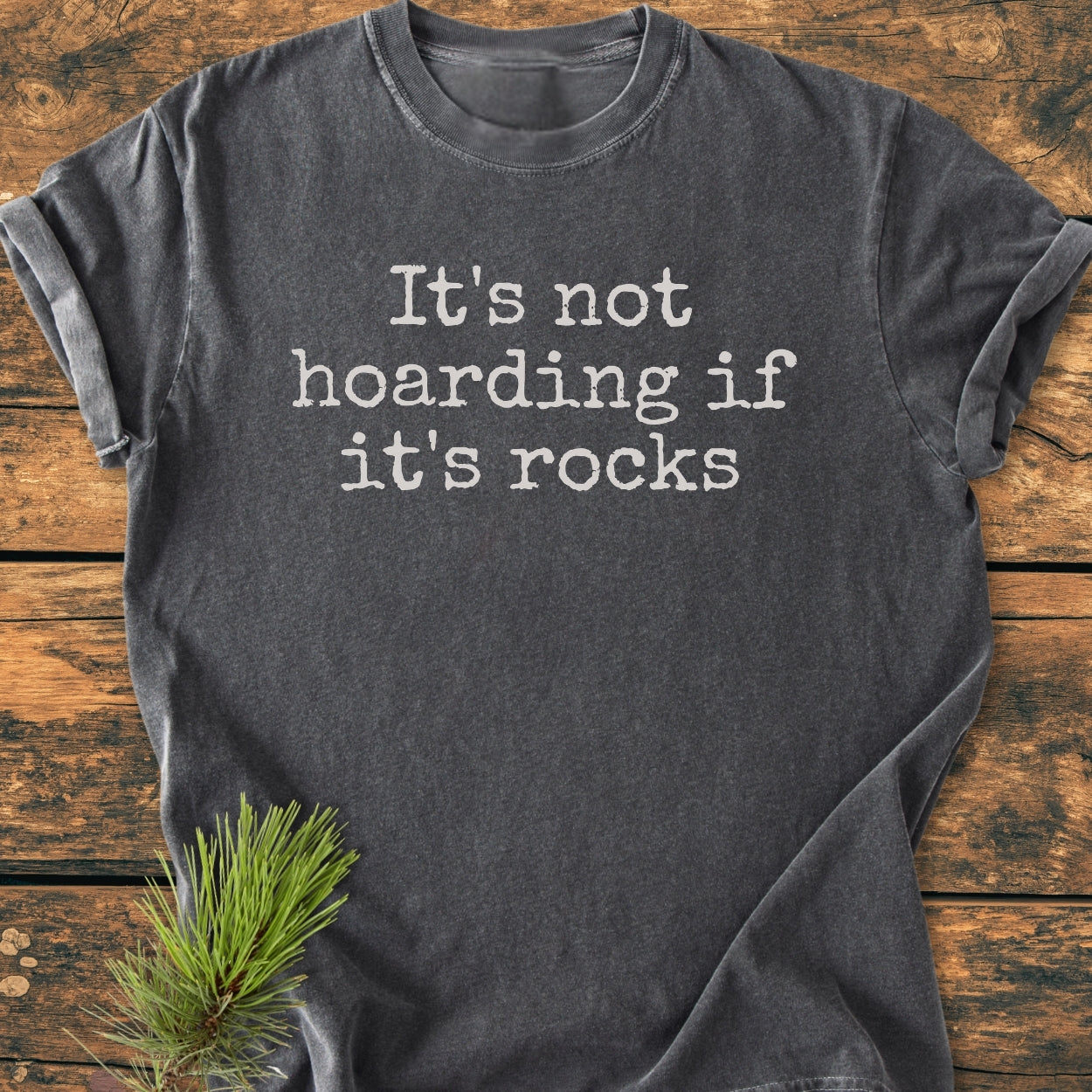 It's Not Hoarding If It's Rocks - Vintage Wash Tee displayed flat with a pine sprig accent. Features white typewriter-style slogan on dark grey fabric, highlighting the relaxed fit and soft-washed texture.