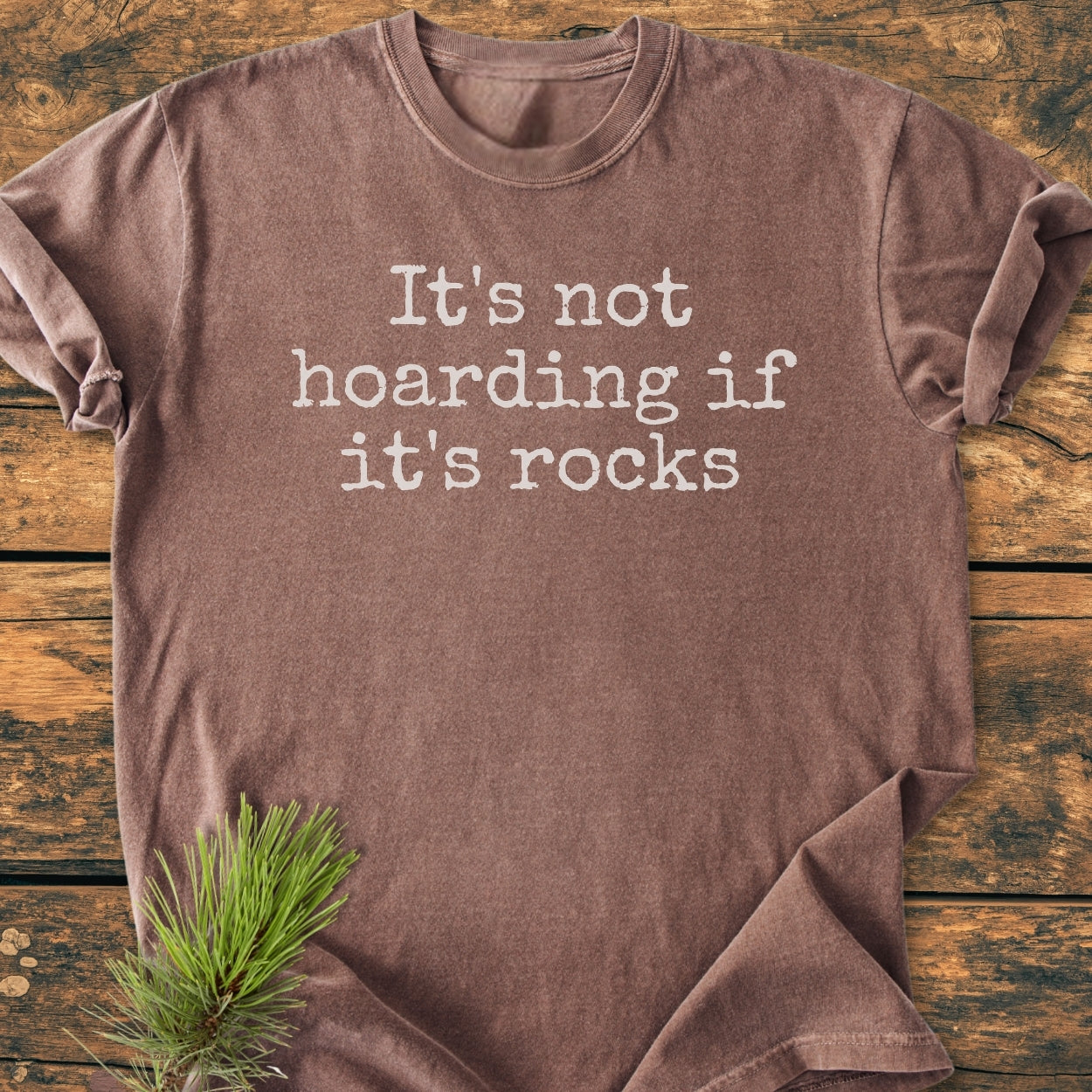The It's Not Hoarding If It's Rocks - Vintage Wash Tee is displayed flat, showcasing its typewriter-style text. A pine branch accents the scene, emphasizing the shirt's relaxed, cozy design.