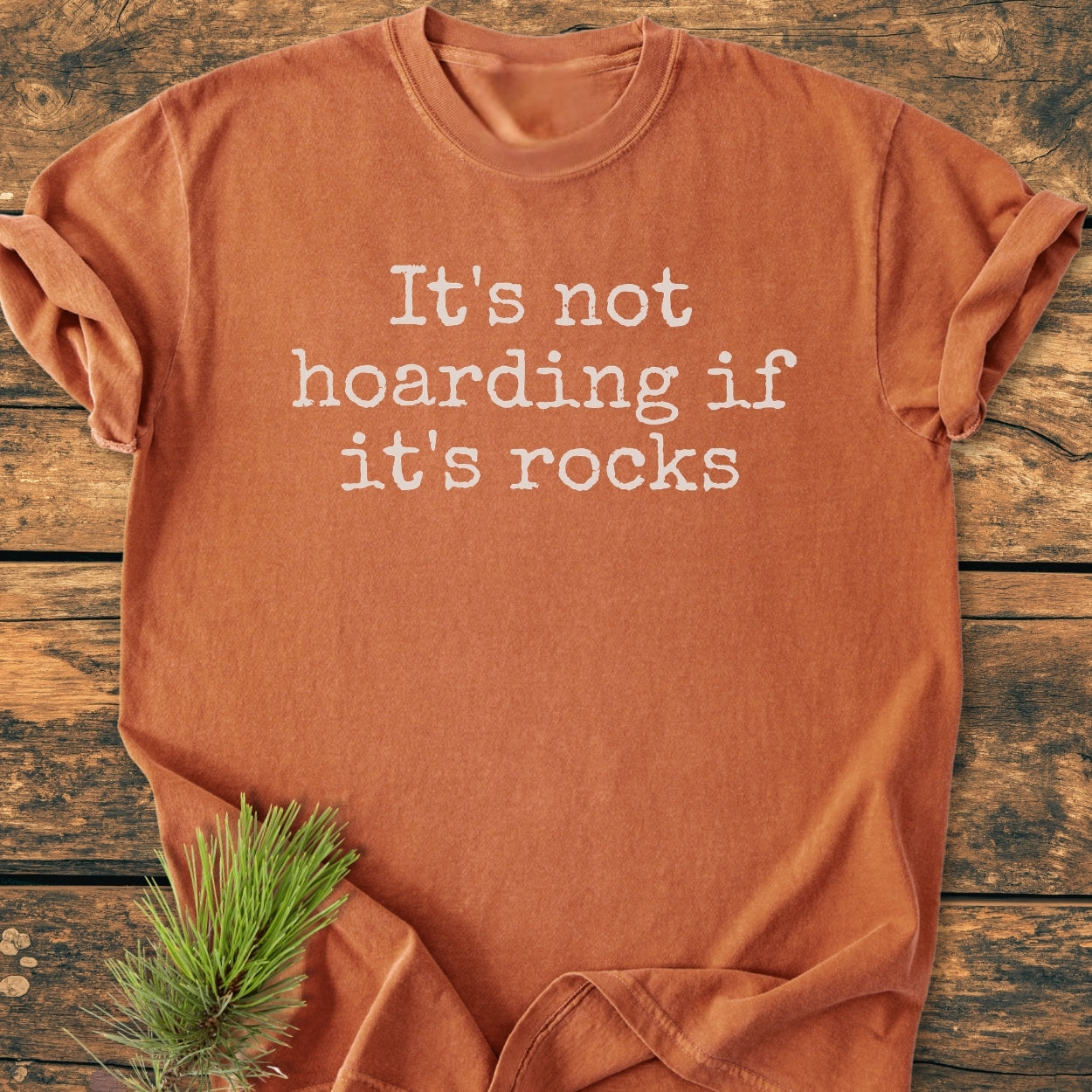 It's Not Hoarding If It's Rocks - Vintage Wash Tee: A muted rust-orange T-shirt with white typewriter-style text, laid on wood with pine sprig. Soft, durable, relaxed fit, 100% cotton.