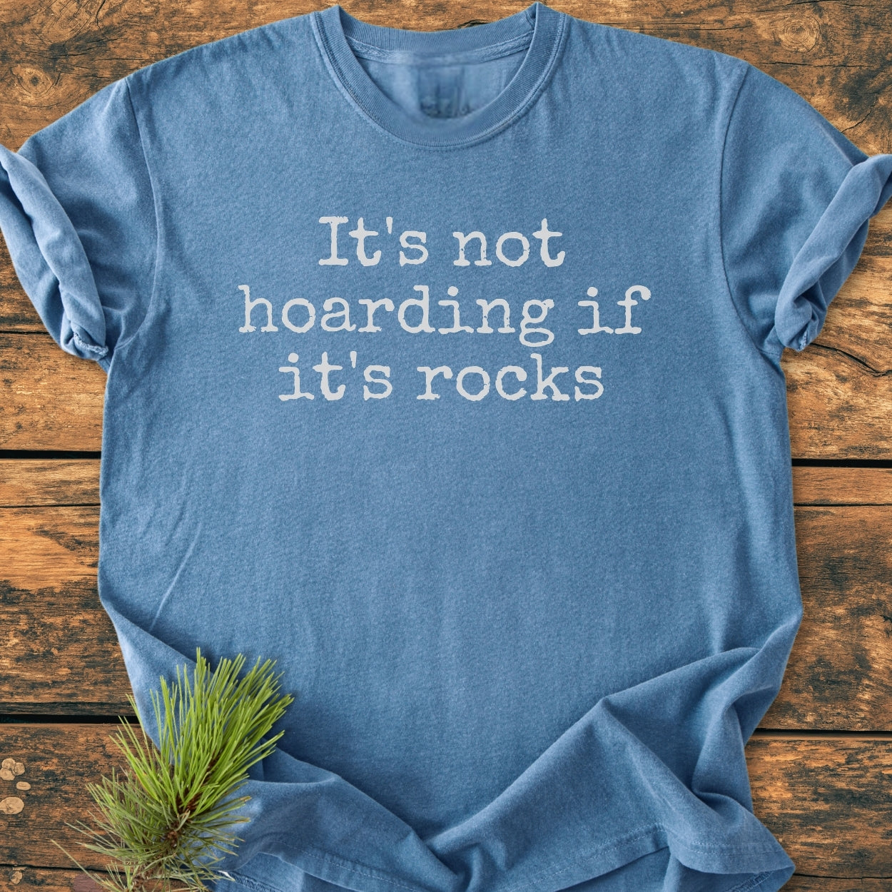 It's Not Hoarding If It's Rocks - Vintage Wash Tee in heather-blue, featuring white typewriter-style text, laid flat with pine sprig accent, showcasing the tee's comfortable, relaxed fit and durable construction.