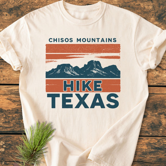 Chisos Mountains Texas - Vintage Wash Tee with a stylized mountain graphic in rusty orange and deep teal, laid on wood, highlighting HIKE TEXAS text; includes a pine sprig accent.