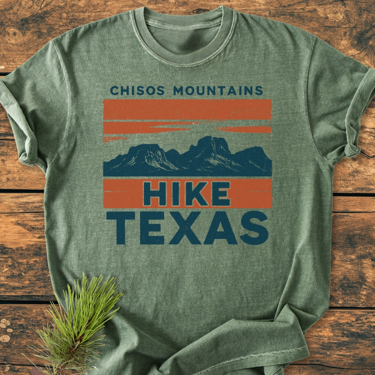 Chisos Mountains Texas - Vintage Wash Tee, featuring a stylized mountain range graphic and “HIKE TEXAS” text. Laid on wood, accompanied by a pine sprig, showcasing its relaxed, cozy fit.