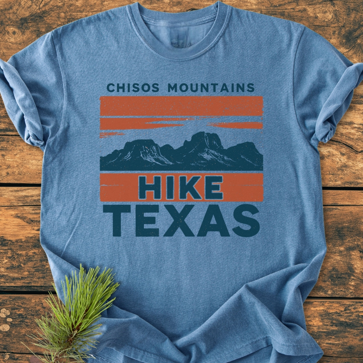 Chisos Mountains Texas - Vintage Wash Tee in heather-blue, featuring a retro “CHISOS MOUNTAINS” print with mountain peaks and “HIKE TEXAS” text, styled with a pine sprig on a wooden surface.